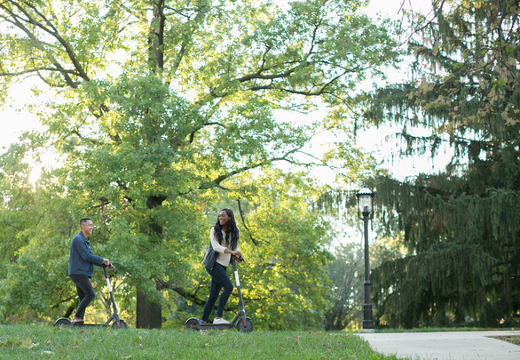 Two people on scooters in park on campus