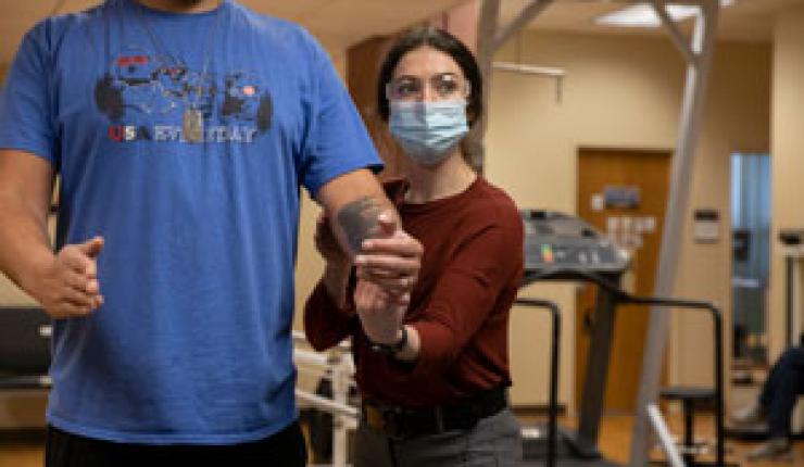 Facing the camera, physical therapist carefully adjusts patient's left arm as he stands and holds both at a 90 degree angle